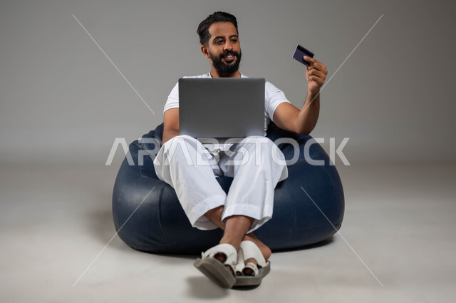 استخدام البطاقة البنكية للدفع الالكتروني، بورتريه لرجل عربي سعودي خليجي مبتسم يرتدي السروال والفنيله البيضاء يجلس في مكان مريح يعمل على جهاز الحاسوب المحمول، إمكانية تسديد الفواتير عبر البرامج الاكترونيه، صورة الجسم بالكامل، خلفية رمادية
