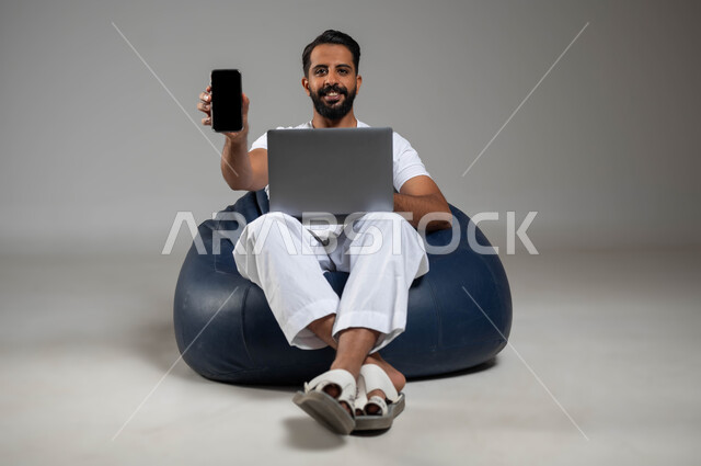 دمج العمل مع التقنية والتكنولوجيا, استخدام تطبيقات الحاسب المحمول الحديثة والمتطورة, إدراة الأعمال عبر الأونلاين, بورتريه لشاب عربي خليجي سعودي يرتدي السوال والفنيلة البيضاء يعرض شاشة الجوال بشاشة سوداء فارغة, صورة الجسم بالكامل, خلفية رمادية