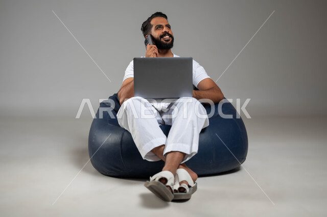 إجراء مكالمة هاتفية عبر الجوال, استخدام الأجهزة والتقنيات الحديثة والمتطورة, بورتريه لشاب عربي خليجي سعودي يرتدي السروال والفنيلة البيضاء يستخدم الحاسب المحمول, إدارة الأعمال عبر الأونلاين, صورة الجسم بالكامل, خلفية رمادية