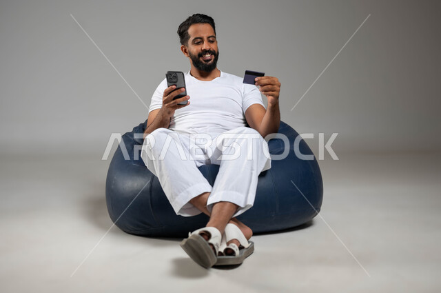 Utilisation d'une carte bancaire pour le paiement électronique, portrait d'un homme souriant du Golfe saoudien portant un pantalon et un T-shirt blanc, assis dans un endroit confortable utilisant un téléphone portable, la possibilité de payer des factures via des programmes électroniques, photo du corps entier, gris arrière-plan