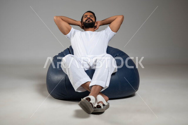 تعابير التأمل والتفكير وشرود الذهن، بورتريه لرجل عربي سعودي خليجي  يرتدي السروال والفانيله البيضاء مستلقي في مكان مريح ينظر إلى شيء ما, إيماءات الراحة والإسترخاء، خلفية رمادية