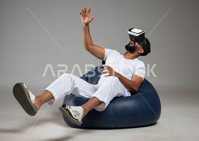 الاندماج والتفاعل مع ألعاب الواقع الافتراضي، مشاهدة الأفلام و محاكاة العالم الخيالي، إيماءات الدهشة والذهول، بورتريه لشاب عربي سعودي خليجي يرتدي السروال والفنيلة البيضاء ونظارة في آر الافتراضية ثلاثية الأبعاد، صورة الجسم بالطول الكامل، خلفية رمادية