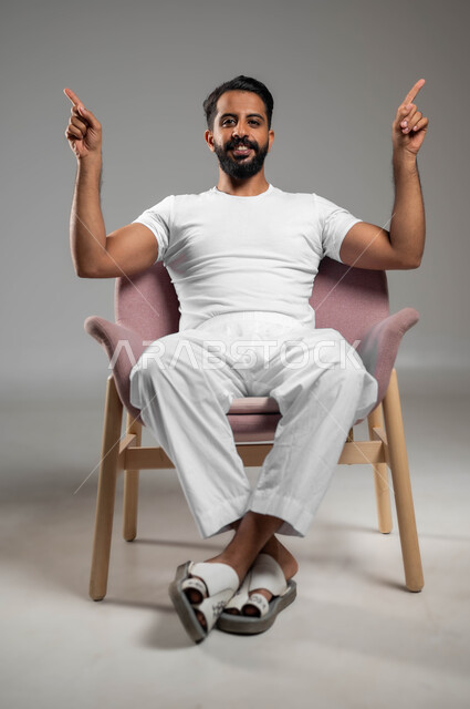 البشاشة والسرور وتحديد شيء معين, رفع اليدين إلى الأعلى والنظر إلى الكاميرا, بورتريه لشاب عربي خليجي سعودي مبتسم يرتدي السروال والفنيلة البيضاء يجلس على الكرسي ويشير بأصابعه السبابة إلى شيء ما, خلفية رمادية