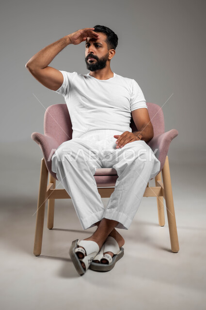 الوقاية من أشعة الشمس, الجلوس والنظر إلى شيء ما, التركيز على رؤية شيء بعيد, بورتريه لشاب عربي خليجي سعودي يرتدي السروال والفنيلة البيضاء يضع يديه على رأسه بإيماءات الدقة, صورة الجسم بالطول الكامل, خلفية رمادية