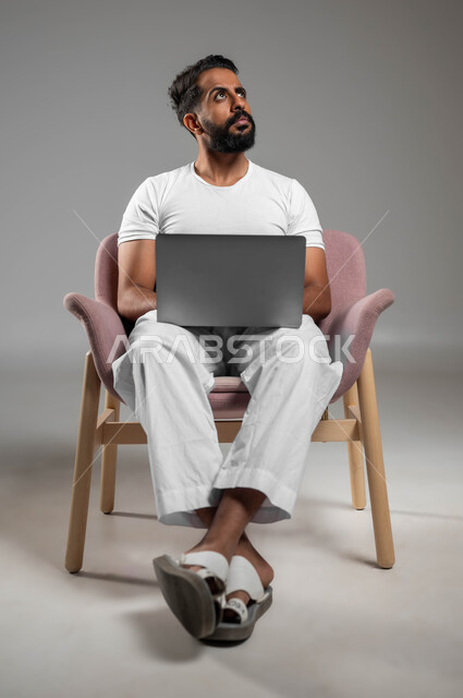 تطور التكنولوجيا والأجهزة التقنية، بورتريه لرجل عربي سعودي خليجي يرتدي السروال والفنيله البيضاء يجلس على كرسي مريح يقوم باستخدام اللابتوب، تعابير التأمل والتفكير، خلفية رمادي