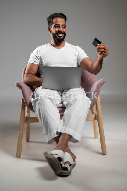 الدفع عبر تطبيقات اللابتوب، استخدام البطاقة البنكية الالكترونية، بورتريه لرجل عربي سعودي خليجي مبتسم يرتدي السروال والفنيله البيضاء يجلس على كرسي مريح يعمل على جهاز الحاسوب المحمول، إمكانية تسديد الفواتير عبر البرامج الكترونيا، صورة لكامل الجسم، خلفية رمادية