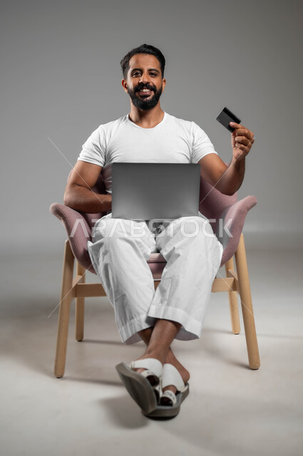 استخدام البطاقة البنكية الالكترونية، بورتريه لرجل عربي سعودي خليجي مبتسم يرتدي السروال والفنيله البيضاء يجلس على كرسي مريح يعمل على جهاز الحاسوب المحمول، الدفع عبر تطبيقات اللابتوب، إمكانية تسديد الفواتير عبر البرامج الكترونيا، صورة لكامل الجسم، خلفية رمادية