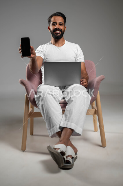 The concept of development and technology, the use of modern and advanced technologies, a portrait of a smiling Saudi Gulf Arab man wearing pants and a T-shirt sitting on a chair holding a mobile phone in his hand and the laptop showing the mobile screen, making phone calls via mobile phone, full body portrait, gray background