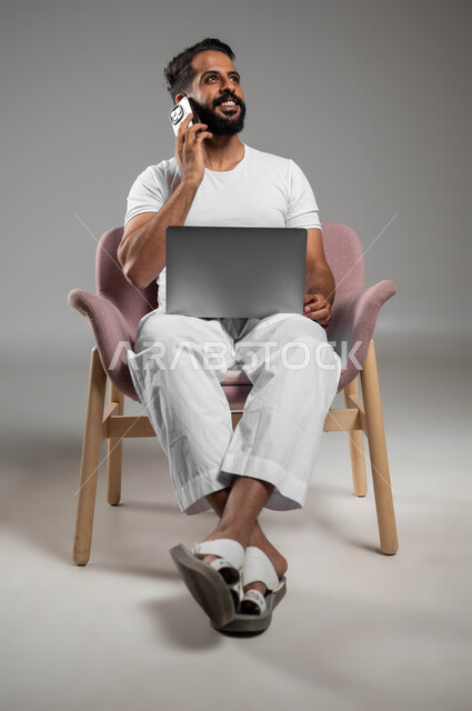 التواصل مع الأهل والأصدقاء، إجراء مكالمات هاتفية عبر الجوال، بورتريه مقرب لرجل عربي سعودي خليجي مبتسم يرتدي السروال والفنيله البيضاء يحمل بيده الهاتف المحمول واللابتوب، مفهوم التطور والتكنولوجيا الحديثة، استخدام الأجهزة والتقنيات المتطورة، خلفية رمادية