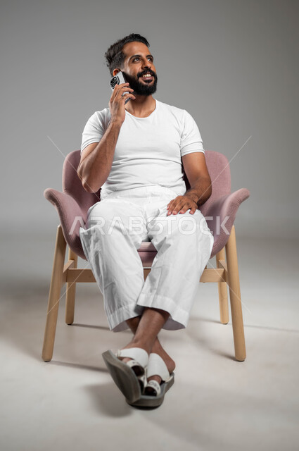 إجراء مكالمات هاتفية عبر الجوال، بورتريه لرجل عربي سعودي خليجي مبتسم يرتدي السروال والفنيله البيضاء يحمل بيده الهاتف المحمول، استخدام الأجهزة والتقنيات المتطورة، التواصل مع الأهل والأصدقاء، صورة الجسم بالطول الكامل، خلفية رمادية