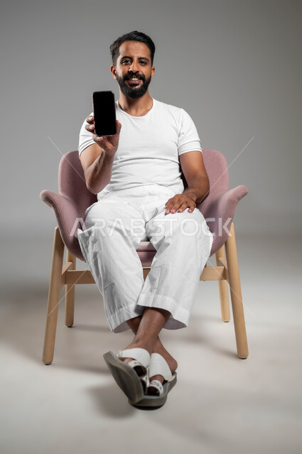 النظر إلى الكاميرا بإيماءات السعادة والسرور، مفهوم التطور والتكنولوجيا، استخدام التقنيات الحديثة والمتطورة، بورتريه لرجل عربي خليجي سعودي مبتسم برتدي سروال وفنيله يجلس على كرسي مريح يحمل بيده الهاتف المحمول يقوم بعرض شاشة الموبايل، صورة لكامل الجسم، خلفية رمادية