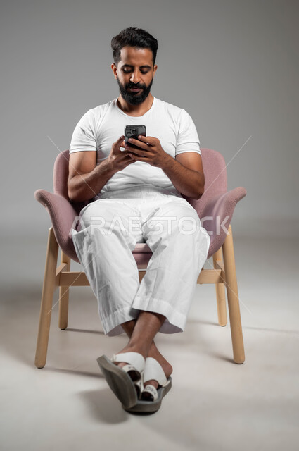 Online shopping, communicating with family and friends, browsing mobile programs and applications, a portrait of a Saudi Gulf Arab man wearing pants and a white T-shirt, holding a mobile phone in his hand, using advanced devices and technologies, a full-body portrait, gray background