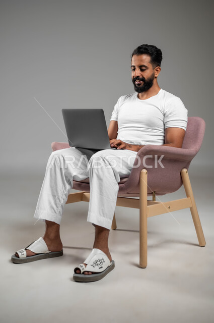 انجاز الأعمال من المنزل عبر الانترنت، مفهوم العمل عن بعد، تصفح برامج و تطبيقات الابتوب، بورتريه  لرجل عربي سعودي خليجي يرتدي السروال والفنيله البيضاء يجلس على كرسي  يقوم باستخدام اللابتوب، الأجهزة والتقنيات المتطورة، صورة لكامل الجسم، خلفية رمادية