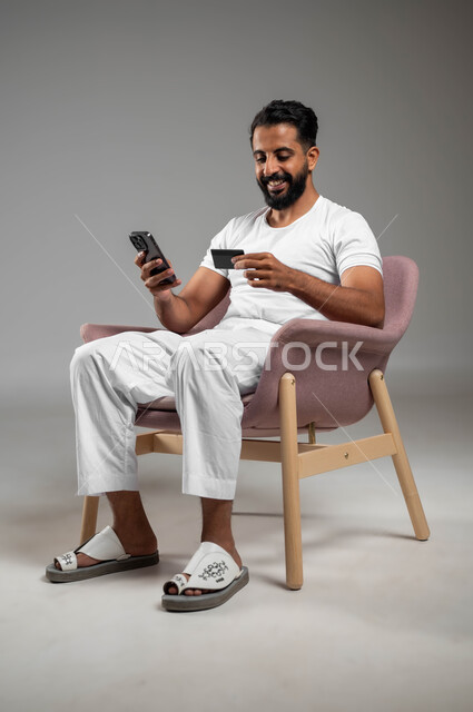 مفهوم الدفع والتسديد عن بعد عبر تطبيقات الجوال، التسوق الالكتروني عبر الانترنت، استخدام البطاقة البنكية الالكترونية، بورتريه لرجل عربي سعودي خليجي مبتسم يرتدي السروال والفنيله البيضاء يجلس على كرسي مريح يحمل الهاتف المحمول، خلفية رمادية