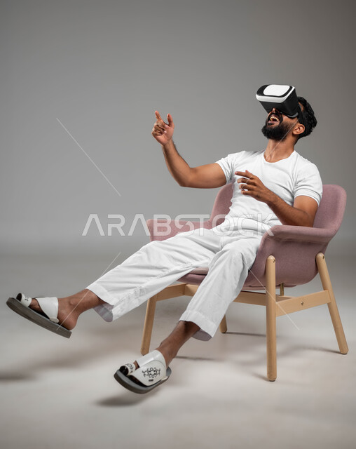 إيماءات التفاعل والاندماج مع العناصر الهولوجرامية، مفهوم التقنيات الإلكترونية الحديثة، بورتريه لرجل عربي سعودي خليجي يجلس على كرسي مريح يرتدي السروال والفنيله البيضاء ونظارات الواقع الإفتراضي (في آر) ثلاثية الأبعاد، خلفية رمادية