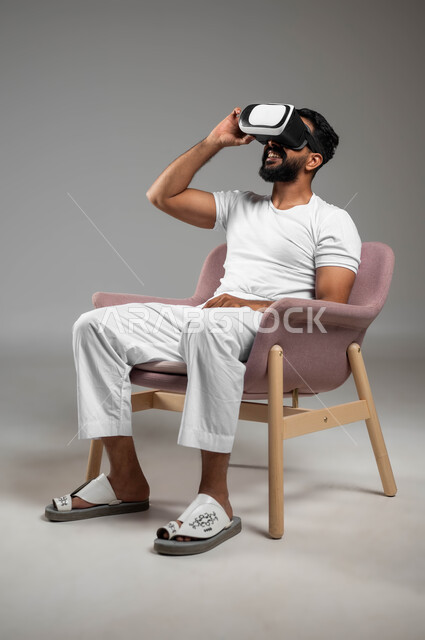 مفهوم التقنيات الإلكترونية الحديثة، إيماءات التفاعل والاندماج مع العناصر الهولوجرامية، بورتريه لرجل عربي سعودي خليجي يجلس على كرسي مريح يرتدي السروال والفنيله البيضاء ونظارات الواقع الإفتراضي (في آر) ثلاثية الأبعاد، خلفية رمادية