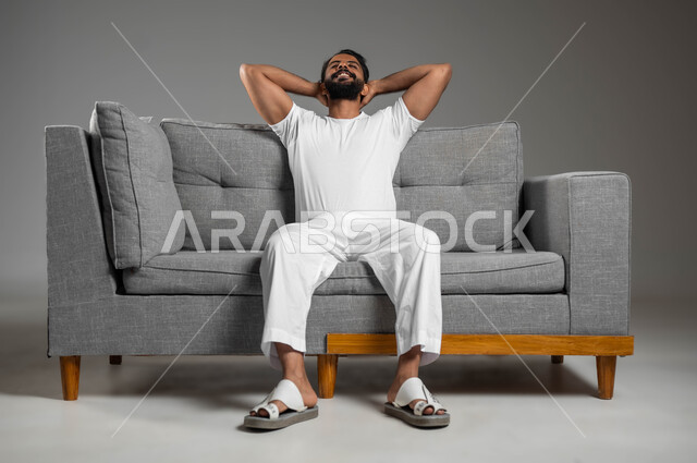 Gestures of rest and relaxation, feeling tired and lethargic and needing sleep, portrait of a Saudi Gulf Arab man wearing pants and a white shirt looking at something sitting on a comfortable sofa, expressions of contemplation and thought, gray background