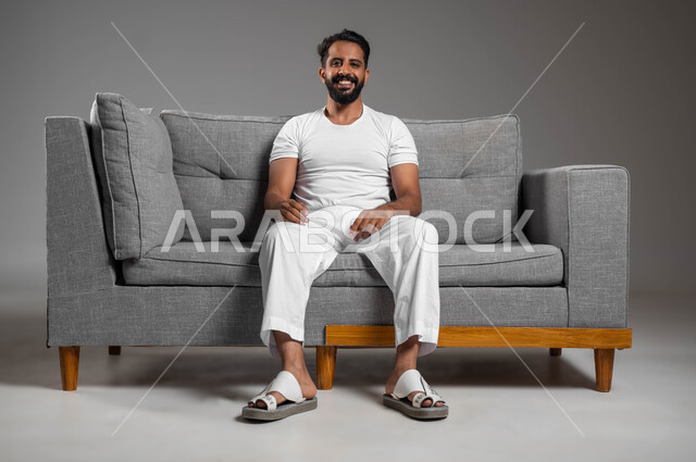 إيماءات السعادة والسرور، , إيماءات الراحة والإسترخاء، بورتريه لرجل عربي سعودي خليجي مبتسم يرتدي السروال والفانيله البيضاء ينظر إلى الكاميرا يجلس على كنبه مريحة, تعابير التأمل والتفكير، خلفية رمادية