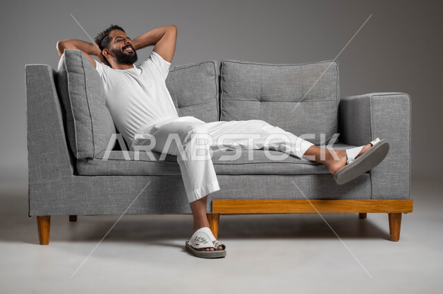 الشعور بالتعب و الخمول والحاجة إلى النوم, إيماءات الراحة والإسترخاء، بورتريه لرجل عربي سعودي خليجي يرتدي السروال والفانيله البيضاء ينظر إلى شيء ما يستريح على الكنبه, تعابير التأمل والتفكير، خلفية رمادية