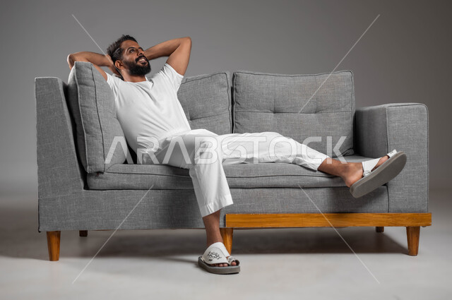 إيماءات الراحة والإسترخاء، الحاجة إلى النوم والشعور بالتعب و الخمول , بورتريه لرجل عربي سعودي خليجي يرتدي السروال والفانيله البيضاء ينظر إلى شيء ما يستريح على الكنبه, تعابير التأمل والتفكير، خلفية رمادية