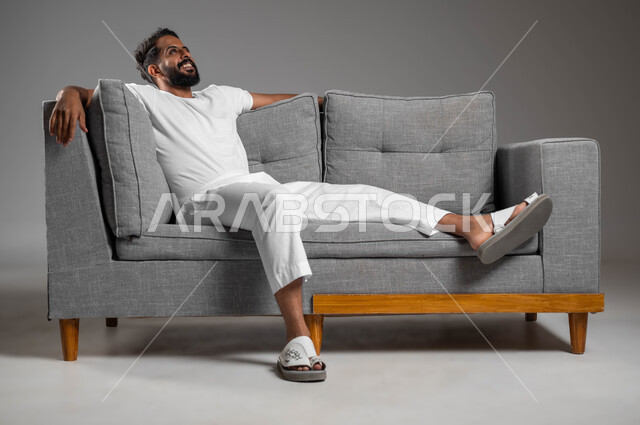 الحاجة إلى النوم والشعور بالتعب و الخمول , إيماءات الراحة والإسترخاء، بورتريه لرجل عربي سعودي خليجي يرتدي السروال والفانيله البيضاء ينظر إلى شيء ما يستريح على الكنبه, تعابير التأمل والتفكير، خلفية رمادية