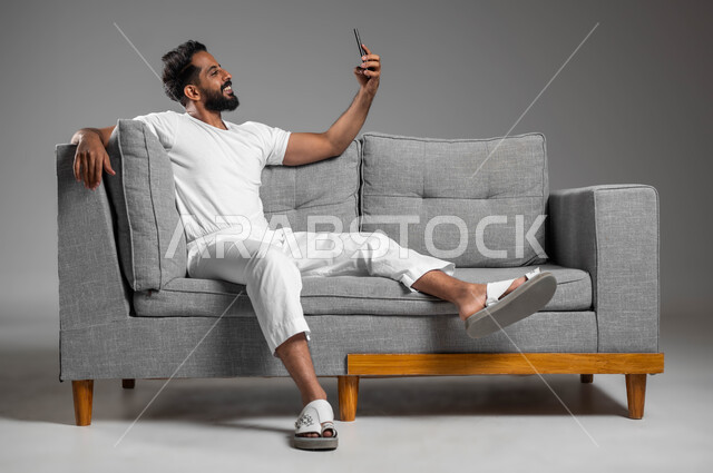 إجراء مكالمة فيديو مع الأهل والأصدقاء، بورتريه لرجل عربي سعودي خليجي مبتسم يرتدي السروال والفنيله البيضاء يجلس على كنبه مريحة يحمل الهاتف المحمول يقوم بالتقاط صور السيلفي التذكارية، تطور التكنولوجيا والأجهزة التقنية، خلفية رمادية