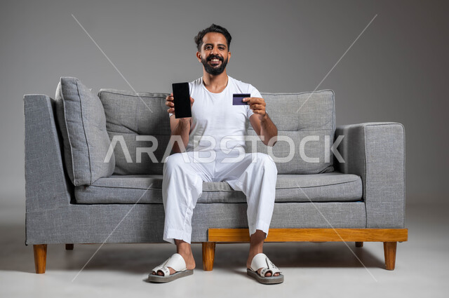 استخدام البطاقة البنكية الالكترونية، مفهوم الدفع والتسديد عن بعد عبر تطبيقات الجوال، بورتريه لرجل عربي سعودي خليجي مبتسم يرتدي السروال والفنيله البيضاء يجلس على كنبه مريحة يقوم بعرض الهاتف المحمول بشاشة سوداء فارغة، خلفية رمادية