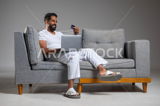 مفهوم الدفع والتسديد عن بعد عبر تطبيقات اللابتوب، استخدام البطاقة البنكية الالكترونية، تطور التكنولوجيا والأجهزة التقنية، بورتريه لرجل عربي سعودي خليجي مبتسم يرتدي السروال والفنيله البيضاء يجلس على كنبه مريحة يعمل على جهاز الحاسوب المحمول، خلفية رمادية