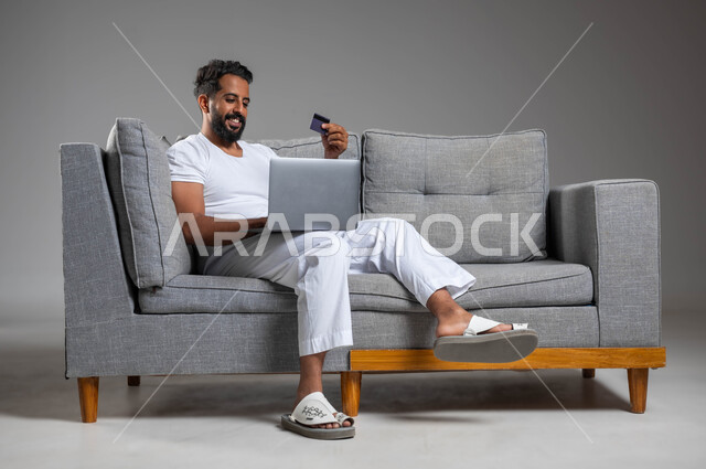 استخدام البطاقة البنكية الالكترونية، تطور التكنولوجيا والأجهزة التقنية الحديثة، مفهوم الدفع والتسديد عن بعد عبر تطبيقات الحاسوب المحمول، بورتريه لرجل عربي سعودي خليجي مبتسم يرتدي السروال والفنيله البيضاء يجلس على كنبه مريحة يعمل على اللابتوب، خلفية رمادية