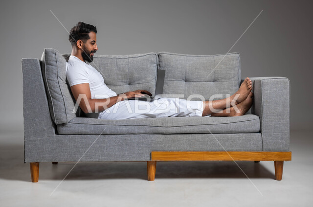 تطور التكنولوجيا والأجهزة التقنية الحديثة، مفهوم العمل عن بعد عبر جهاز الحاسوب المحمول، بورتريه لرجل عربي سعودي خليجي مبتسم يرتدي السروال والفنيله البيضاء يجلس على كنبه مريحة يستخدم  اللابتوب، خلفية رمادية