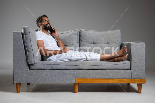 إجراء مكالمة هاتفية مع الأهل والأصدقاء، بورتريه لرجل عربي سعودي خليجي مبتسم يرتدي السروال والفنيله البيضاء يجلس على كنبه مريحة يحمل الهاتف المحمول، تطور التكنولوجيا والأجهزة التقنية، إيماءات الفرح والسرور، خلفية رمادية