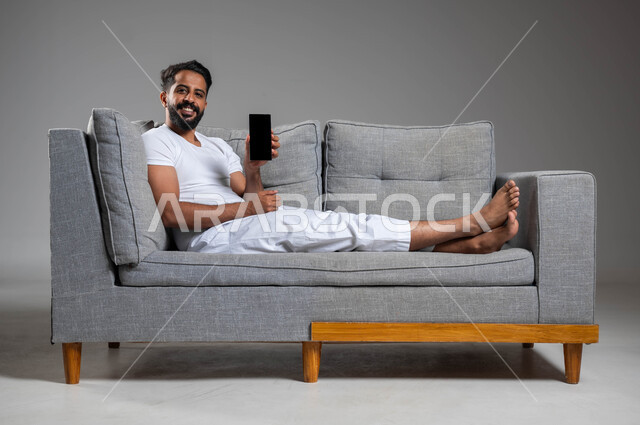 تطور التكنولوجيا والأجهزة التقنية الحديثة، بورتريه لرجل عربي سعودي خليجي مبتسم يرتدي السروال والفنيله البيضاء ينظر إلى الكاميرا يجلس على كنبه مريحة يقوم بعرض شاشة سوداء فارغة على الهاتف المحمول، خلفية رمادية