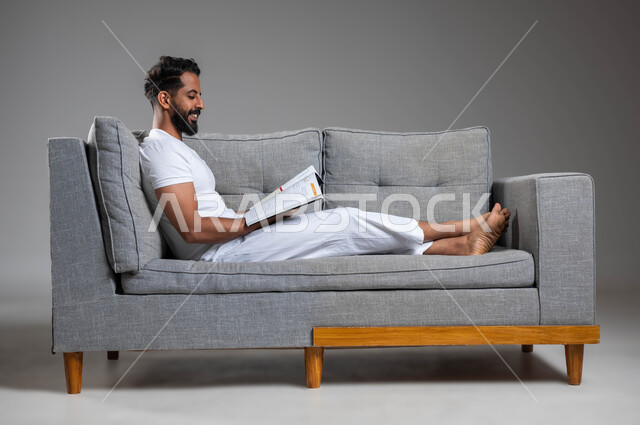 قضاء وقت الفراغ بالتعلم والثقافة، بورتريه لشاب عربي خليجي سعودي مبتسم يرتدي السروال والفنيله البيضاء يستريح على الكنبة يقرأ كتاب, مفهوم التثقيف والبحث العلمي، هواية القراءة والاستكشاف، خلفية رمادية