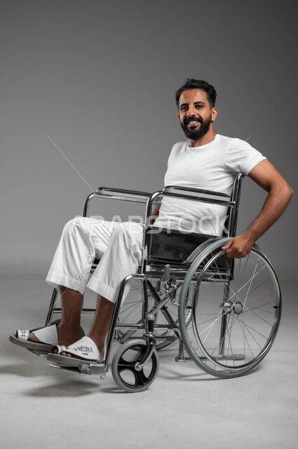 النظر إلى الكاميرا بإيماءات التفاؤل والسرور، أسلوب حياة ذوي الاحتياجات الخاصة، أجهزة ومعدات تساعد على الحركة والتنقل, بورتريه لرجل عربي سعودي خليجي مبتسم يرتدي السروال والفنيله البيضاء يجلس على كرسي متحرك ينظر إلى شيء ما, خلفية رمادية