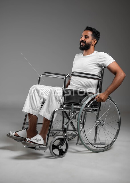 Lifestyle for people with special needs, devices and equipment that help with movement and mobility, portrait of a smiling Saudi Arabian Gulf man wearing pants and a white T-shirt sitting in a wheelchair looking at something, gestures of optimism and pleasure, gray backgroun