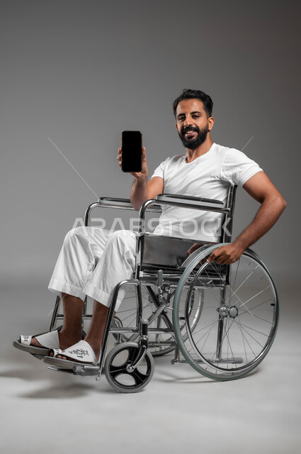 Facilitating the lives of people of determination using technology and technology, a portrait of a smiling Saudi Arabian Gulf man wearing pants and a white T-shirt, sitting in a wheelchair, looking at the camera, holding a mobile phone, displaying a blank black screen, gray background
