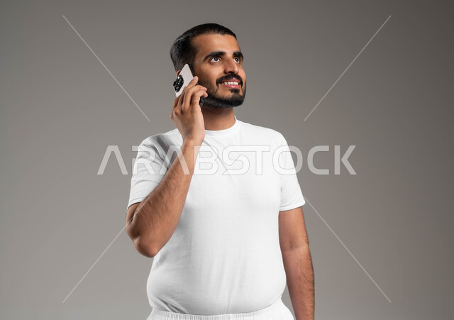 التواصل مع الأهل والأصدقاء، إجراء مكالمات هاتفية عبر الجوال، بورتريه لرجل عربي سعودي خليجي مبتسم يرتدي السروال والفنيله البيضاء يحمل الهاتف المحمول، تطور التكنولوجيا والأجهزة التقنية، خلفية رمادية