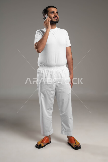 إجراء مكالمات هاتفية عبر الجوال، بورتريه لرجل عربي سعودي خليجي مبتسم يرتدي السروال والفنيله البيضاء يحمل الهاتف المحمول، تطور التكنولوجيا والأجهزة التقنية، التواصل مع الأهل والأصدقاء، صورة الجسم بالطول الكامل، خلفية رمادية