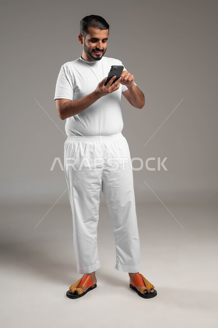 الدردشة مع الأهل والأصدقاء، التسوق الالكتروني وتصفح برامج وتطبيقات الجوال، بورتريه لرجل عربي سعودي خليجي مبتسم يرتدي السروال والفنيله البيضاء يحمل الهاتف المحمول، تطور التكنولوجيا والأجهزة التقنية، صورة الجسم بالطول الكامل، خلفية رمادية