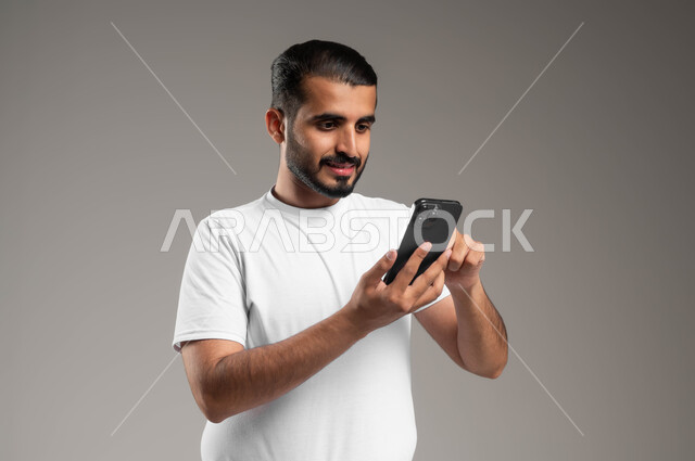 التسوق الالكتروني وتصفح برامج وتطبيقات الجوال، بورتريه لرجل عربي سعودي خليجي مبتسم يرتدي السروال والفنيله البيضاء يحمل الهاتف المحمول، تطور التكنولوجيا والأجهزة التقنية، الدردشة مع الأهل والأصدقاء، خلفية رمادية