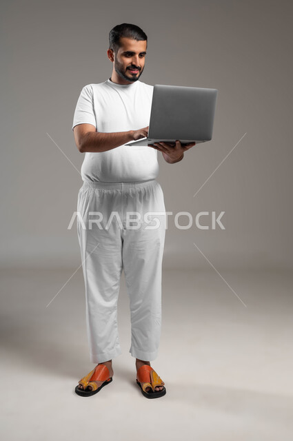 تصفح الانترنت وبرامج التواصل الاجتماعي, إستخدام الحاسوب المحمول لإنجاز المهام، بورتريه لرجل عربي سعودي خليجي مبتسم يرتدي السروال والفنيله البيضاء يحمل جهاز اللابتوب، تطور التكنولوجيا والأجهزة التقنية، صورة الجسم بالطول الكامل، خلفية رمادية