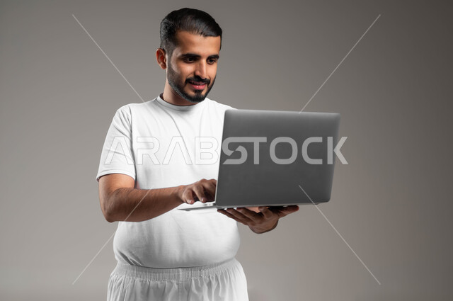 إستخدام الحاسوب المحمول لإنجاز المهام، بورتريه لرجل عربي سعودي خليجي مبتسم يرتدي السروال والفنيله البيضاء يحمل جهاز اللابتوب، تطور التكنولوجيا والأجهزة التقنية الحديثة، تصفح الانترنت وبرامج التواصل الاجتماعي, إجراء مكالمات الفيديو، خلفية رمادية