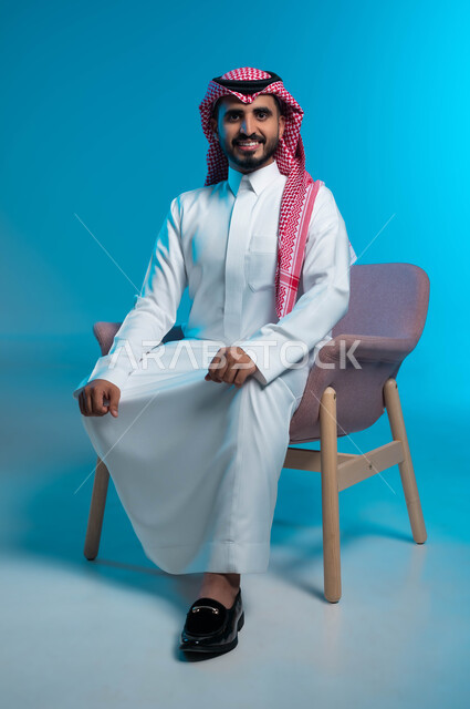 الجلوس بثقة واستقامة, الاهتمام بالمظهر الخارجي والاناقة, بورتريه مقرب لشاب عربي سعودي خليجي يرتدي الثوب والشماغ التقليدي, النظر الى الكاميرا بتمعن وابتسامة, خلفية زرقاء