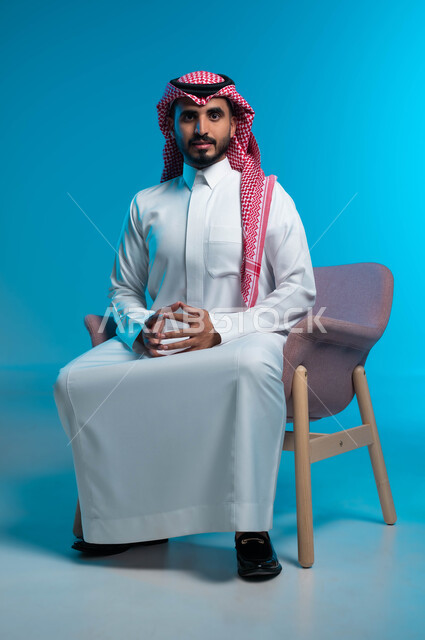 مفهوم الاناقة والاهتمام بالمظهر الخارجي, الجلوس بثقة واستقامة, بورتريه مقرب لشاب عربي سعودي خليجي يرتدي الثوب والشماغ التقليدي يجلس على الكرسي, النظر الى الكاميرا بايماءات القوة, خلفية زرقاء