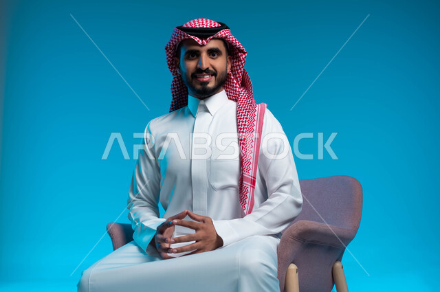 الجلوس بثقة واستقامة, مفهوم الاناقة والاهتمام بالمظهر الخارجي, بورتريه مقرب لشاب عربي سعودي خليجي يرتدي الثوب والشماغ التقليدي يجلس على الكرسي, ايماءات سعادة وسرور وابتسامة, خلفية زرقاء
