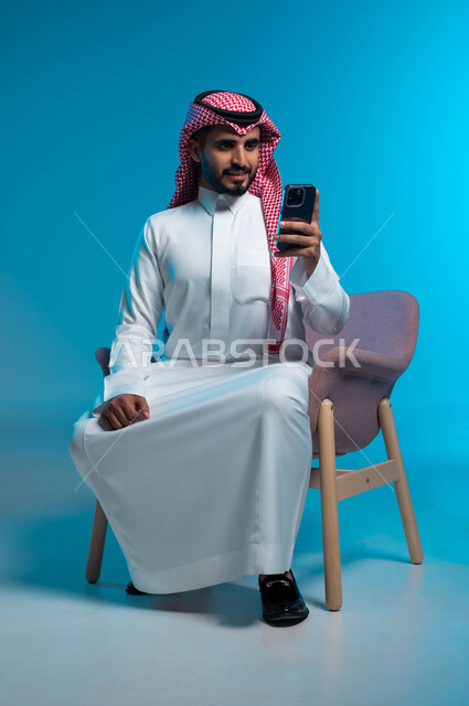 متابعة اخر اخبار مواقع التواصل الاجتماعي, تصفح مواقع الانترنت عبر الهاتف المحمول, الدردشة مع الاصدقاء والاهل, بورتريه لشاب عربي سعودي خليجي يرتدي الثوب والشماغ التقليدي يجلس على الكرسي يمسك بيده الجوال, خلفية زرقاء