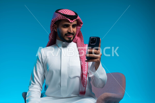 تصفح مواقع الانترنت عبر الهاتف المحمول, الدردشة مع الاصدقاء والاهل, متابعة اخر اخبار مواقع التواصل الاجتماعي, بورتريه مقرب لشاب عربي سعودي خليجي يرتدي الثوب والشماغ التقليدي يجلس على الكرسي يمسك بيده الجوال, خلفية زرقاء