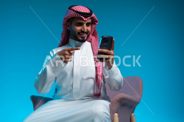 استخدام التكنولوجيا المتطورة, الاشارة الى الهاتف المحمول, بورتريه مقرب لشاب عربي سعودي خليجي يرتدي الثوب والشماغ التقليدي يجلس على الكرسي يمسك بيده الجوال, اجراء مكالمة فيديو مع الاهل والاصدقاء, تعابير الفرح والسعادة في سماع الاخبار الجيدة, خلفية زرقاء