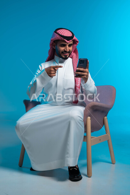 الاشارة الى الهاتف المحمول, استخدام التكنولوجيا المتطورة, بورتريه لشاب عربي سعودي خليجي يرتدي الثوب والشماغ التقليدي يجلس على الكرسي يمسك بيده الجوال, اجراء مكالمة فيديو مع الاهل والاصدقاء, تعابير الفرح والسعادة في سماع الاخبار الجيدة, خلفية زرقاء