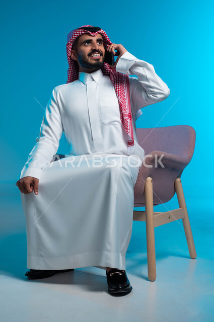Joy and happiness in hearing the good news, making a phone call via mobile phone, chats and work conversations with colleagues, portrait of a smiling Saudi Arabian Gulf young man wearing the traditional thobe and shemagh, holding the mobile phone in his hand, sitting on the chair, blue background